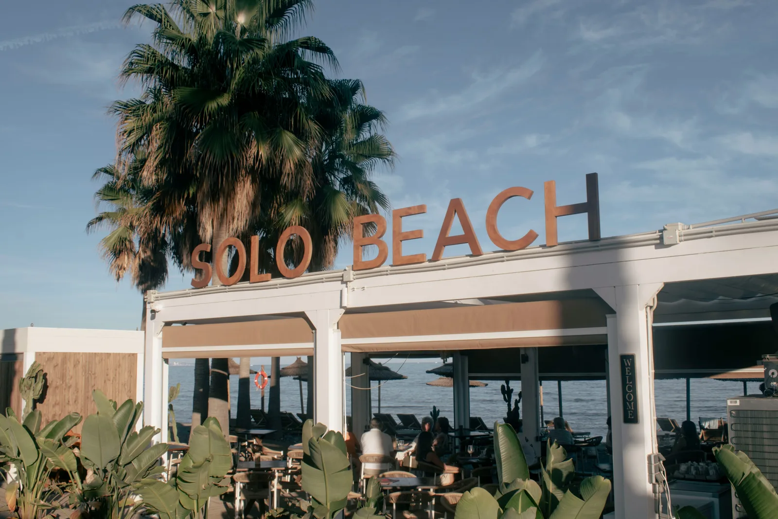 Fachada de Solo Beach con el cartel de letras, palmeras y la terraza frente al mar